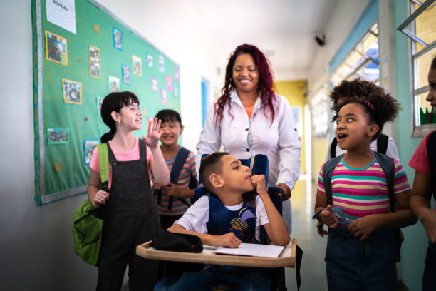 La imagen muestra una escena escolar muy cálida e inclusiva en un pasillo de escuela.

En el centro aparece una maestra sonriendo, rodeada por varios niños que también se ven contentos y conversando entre sí. Al frente hay un niño usando silla de ruedas con una libreta en su mesa; está participando junto con sus compañeros. A su alrededor, otros niños con mochilas lo acompañan y platican animadamente, como si fueran camino al aula o saliendo de clase.

El ambiente transmite **alegría, convivencia y apoyo**, y sugiere una escuela donde todos los estudiantes participan juntos, sin importar sus diferencias.
